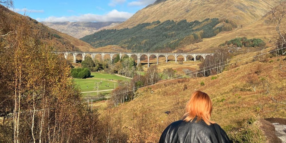 Person with red hair overlooking scenic viaduct in hilly landscape.