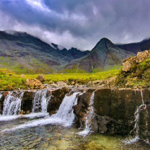 Small waterfall with rocky landscape and mountains under cloudy sky.