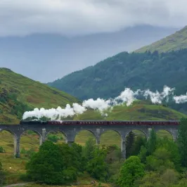 Steam train crossing a stone viaduct in a lush, mountainous landscape.