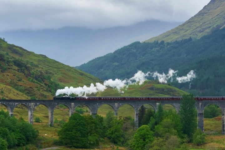 Steam train crossing a stone viaduct in a lush, mountainous landscape.