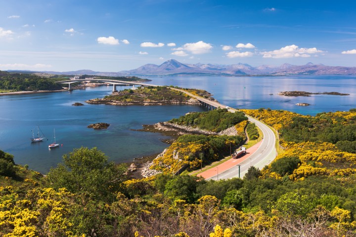 Scenic view of a coastal road, bridge, and mountains with boats and greenery.