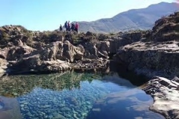 Group of people on rocky terrain near clear water and mountains.