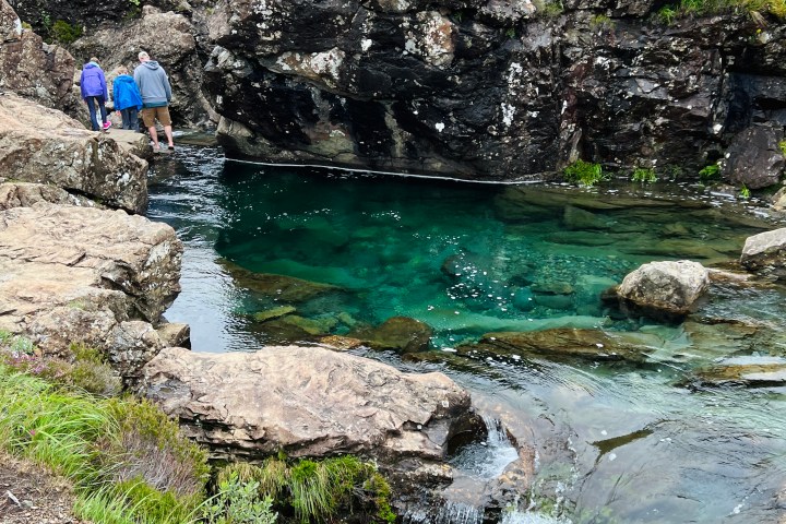People standing by a clear blue pool with rocky cliffs and a small waterfall in a grassy landscape.
