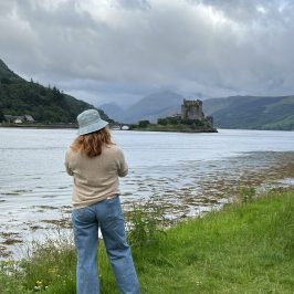 Person in hat by a lake with castle in the distance on a cloudy day.