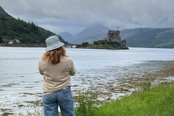 Person in hat by a lake with castle in the distance on a cloudy day.