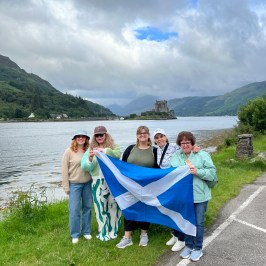 Five people holding Scottish flag by a lake with a castle in the distance.