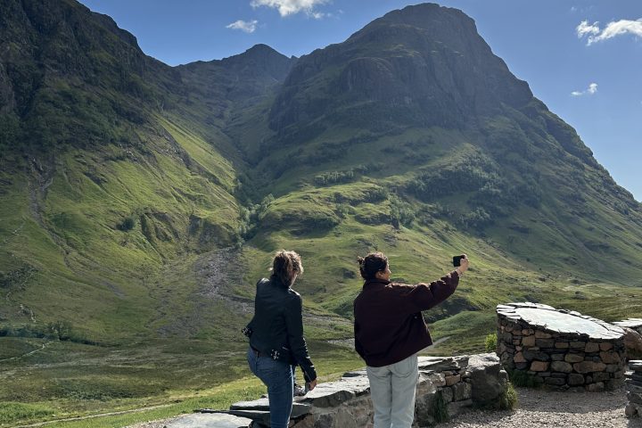 Two people stand on a rocky path, taking photos of a large mountain under a blue sky with clouds.