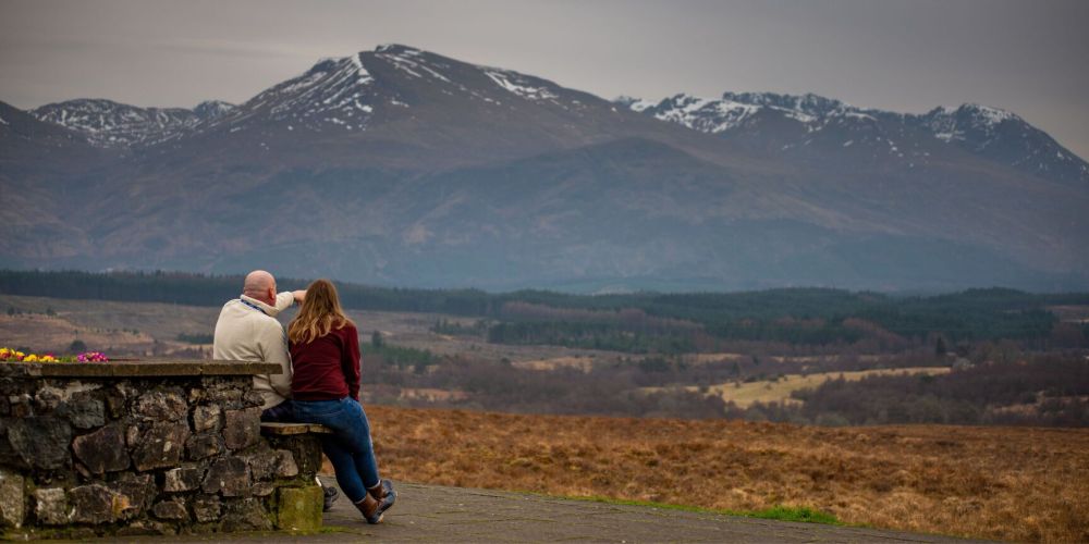 Two people sit on a bench overlooking a mountainous landscape.