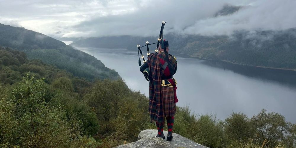 Person in Scottish attire playing bagpipes on a hill overlooking a misty lake and mountains.