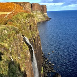 Waterfall cascading down cliffs into the ocean with grassy landscape above.