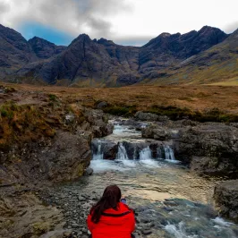 Person in red coat sitting by a stream with mountains in the background.