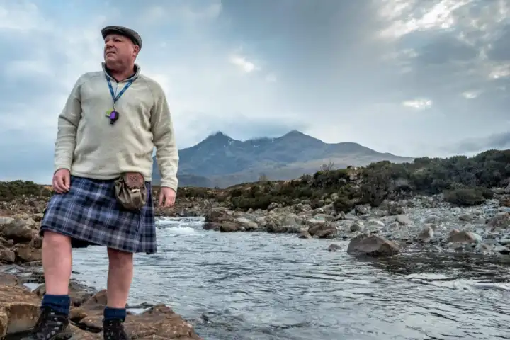 Man in kilt stands by rocky river with mountains and cloudy sky in background.